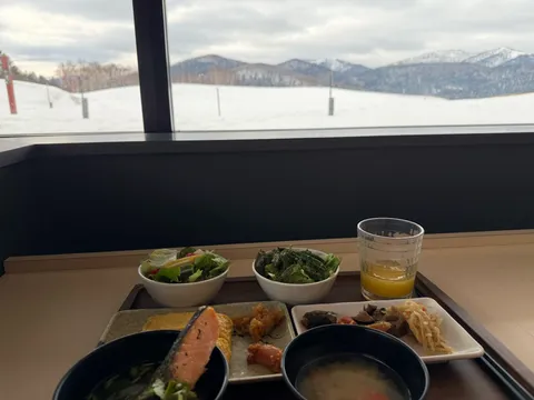 窓の外に雪山を望みながら並ぶ和朝食のトレイ。焼き鮭のお茶漬け・だし巻き・味噌汁・煮物・オレンジジュース