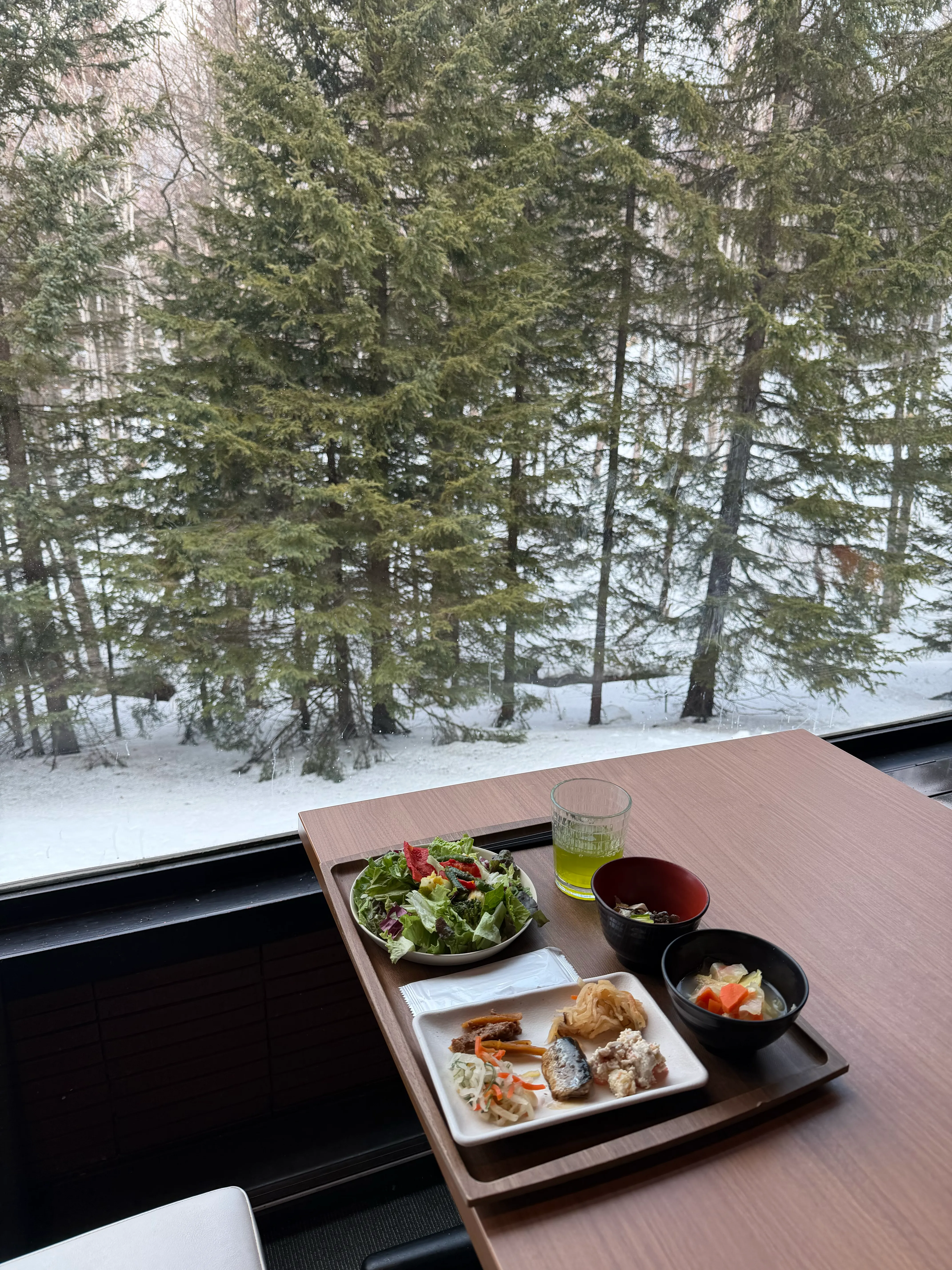 雪景色が広がる窓際テーブルに並ぶ朝食トレイ。サラダ・煮物・味噌汁など和洋ミックスの一皿