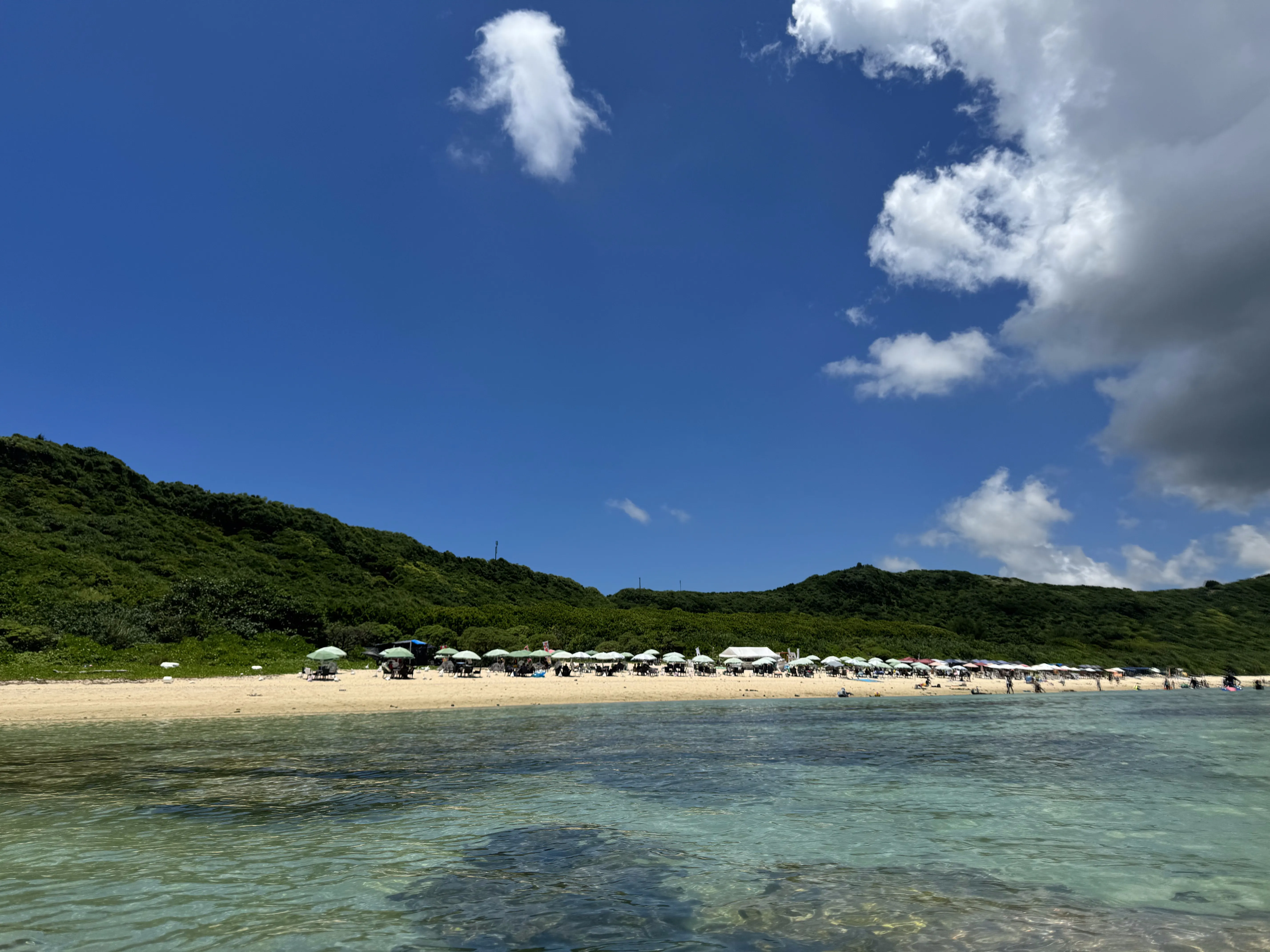 海から望むアラグスク浜。白砂のビーチに色とりどりのパラソルが並び、背後に緑の丘が広がる