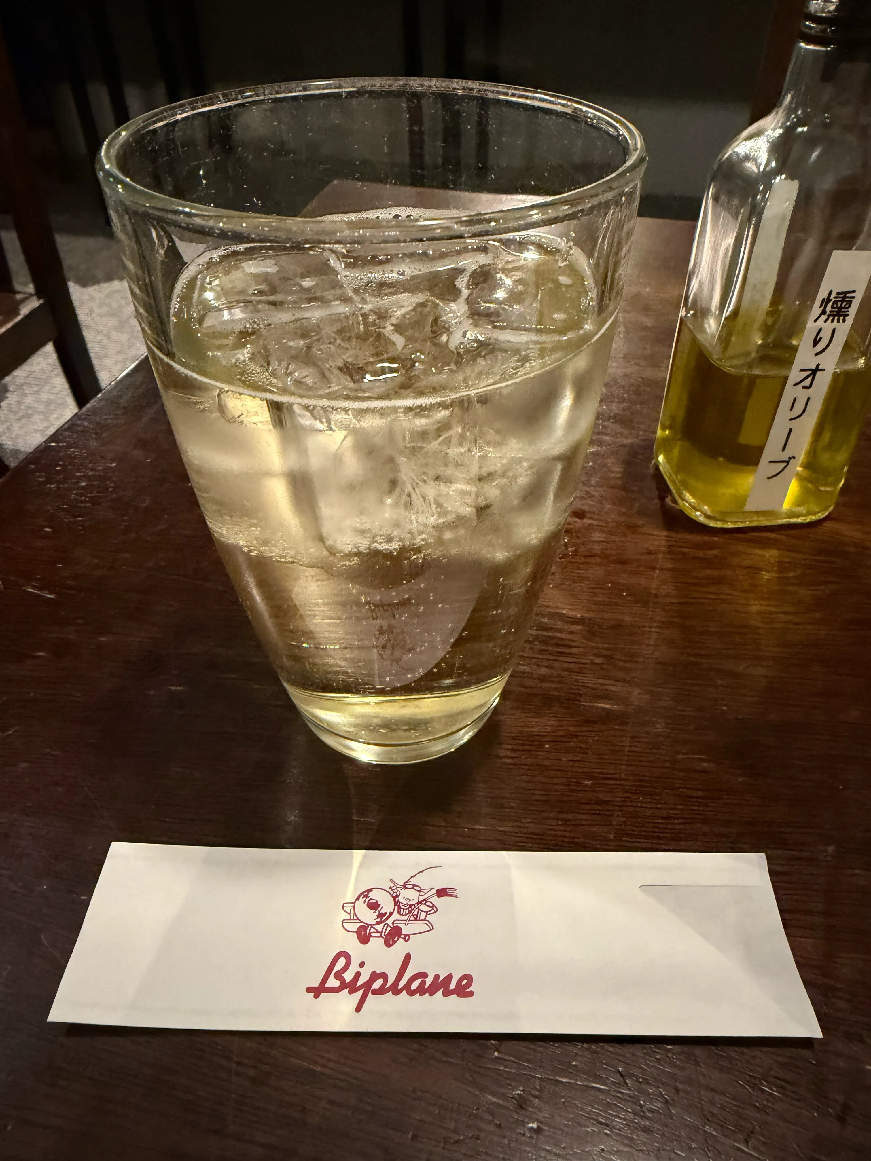 Biplaneのロゴが入ったメニュー封筒とハイボール。氷の入った背の高いグラスとテーブルの上の瓶が夜の時間を演出する
