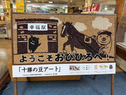 十勝の豆アート。複数種の豆を組み合わせて描かれた「ようこそおびひろへ！」のアート作品。幸福駅と馬とぶたが描かれている