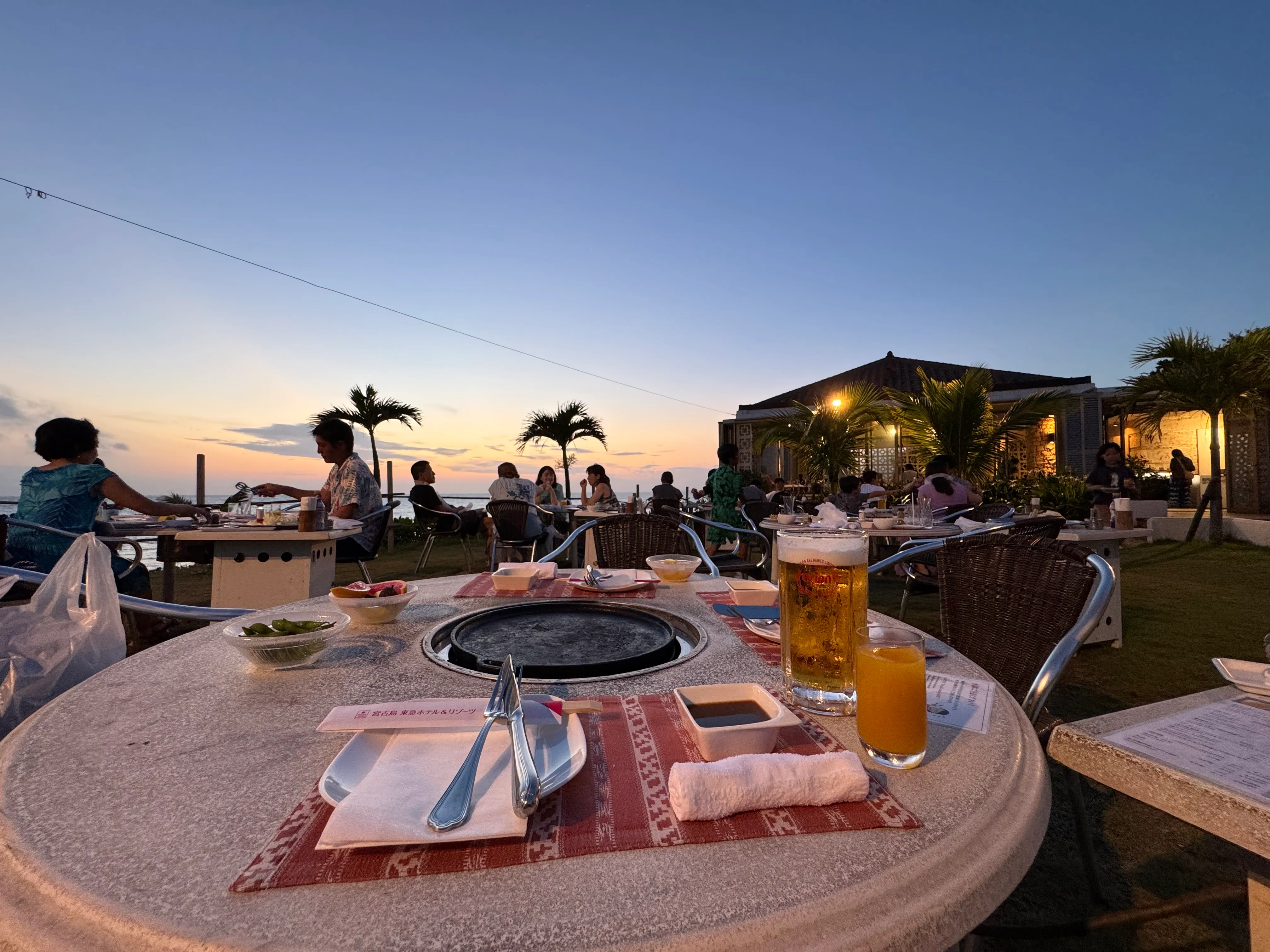 屋外BBQエリアの夕暮れ（ヤシの木シルエット・BBQコンロ・ビールとジュース）