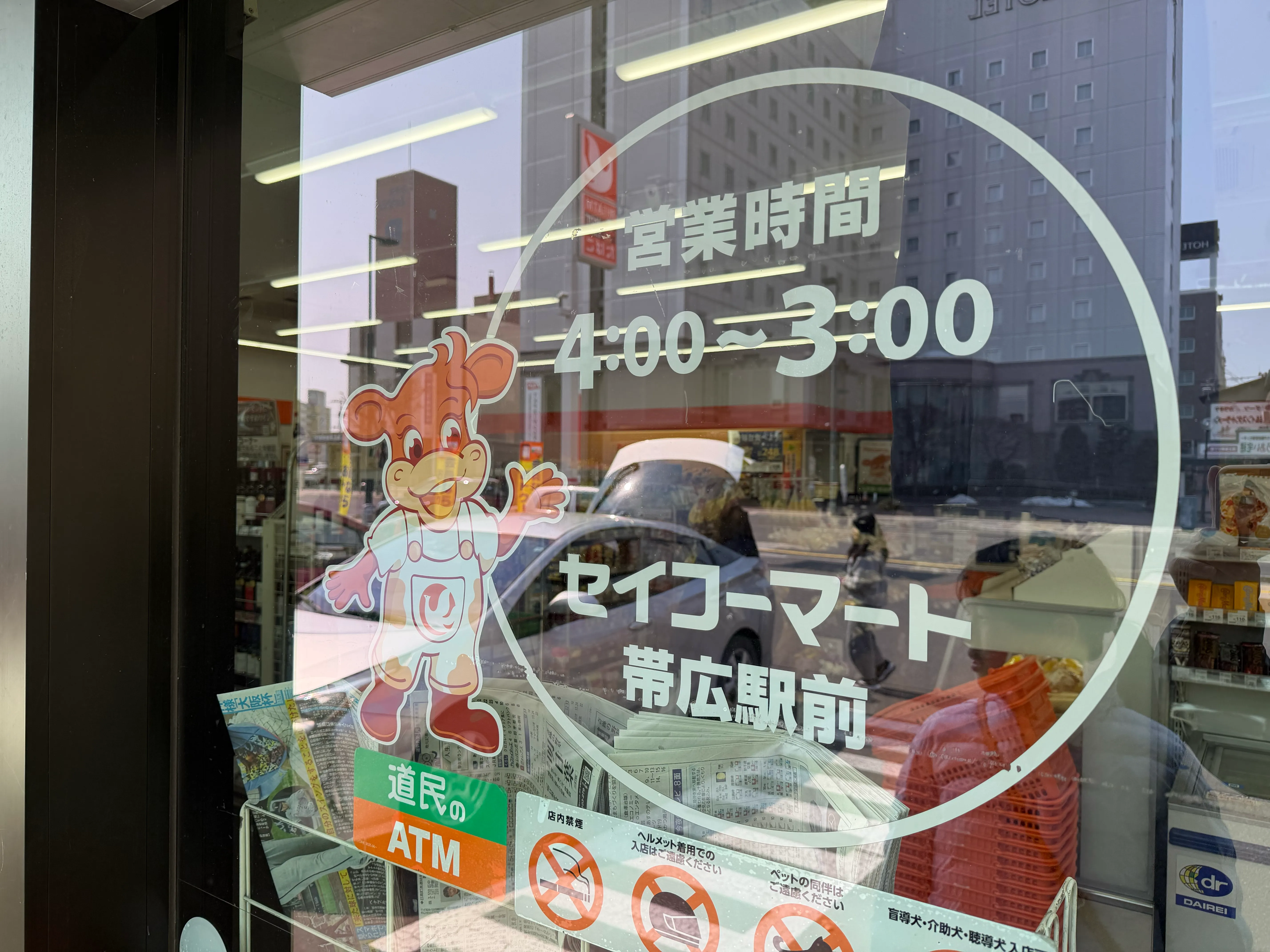 セイコーマート帯広駅前店のガラスドア。営業時間4:00〜3:00と書かれ、道民のATMのステッカーが貼られている