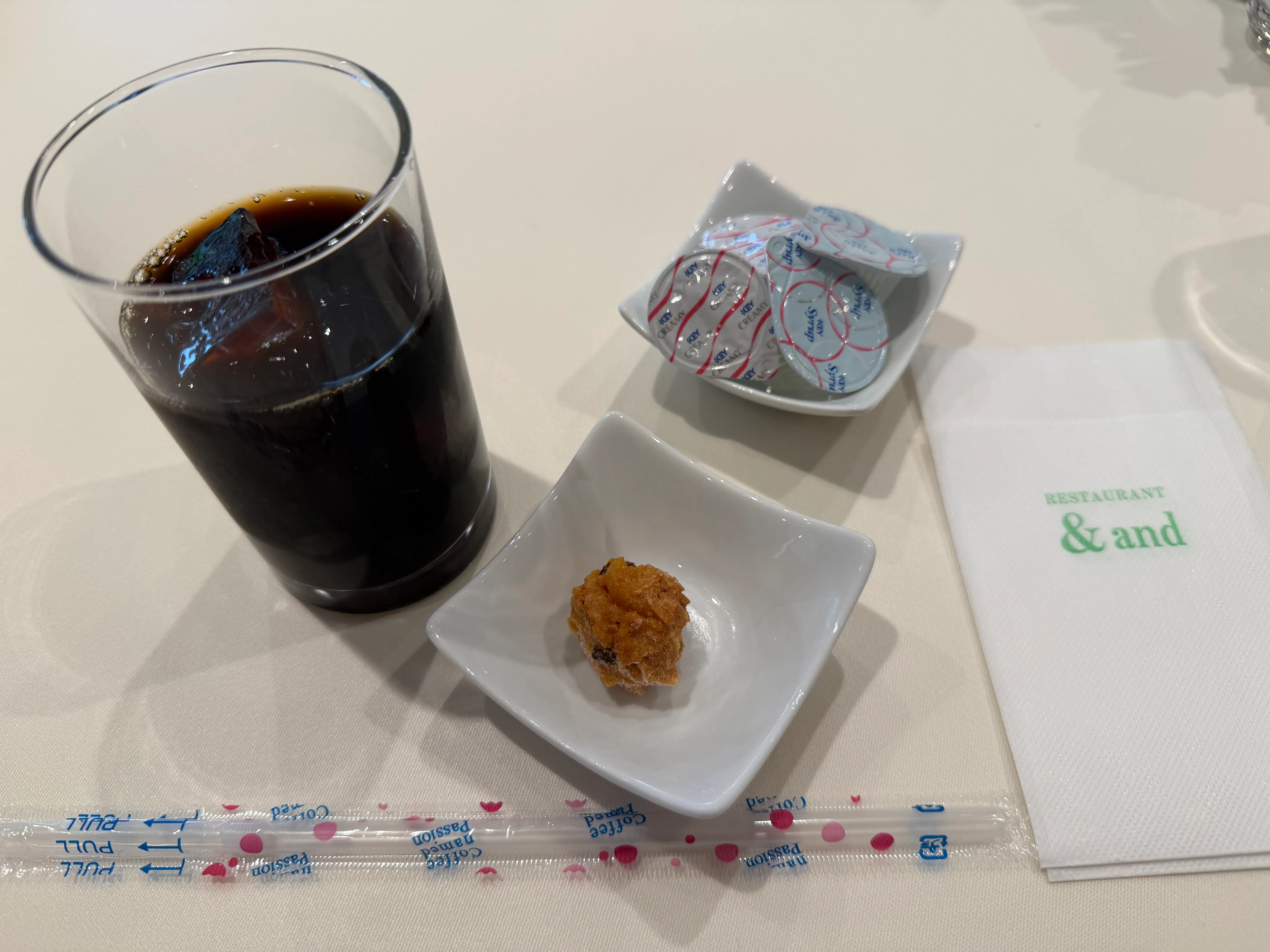 アイスコーヒーと小さな菓子が小皿に。&#x26;andのグリーンのナプキンが添えられている