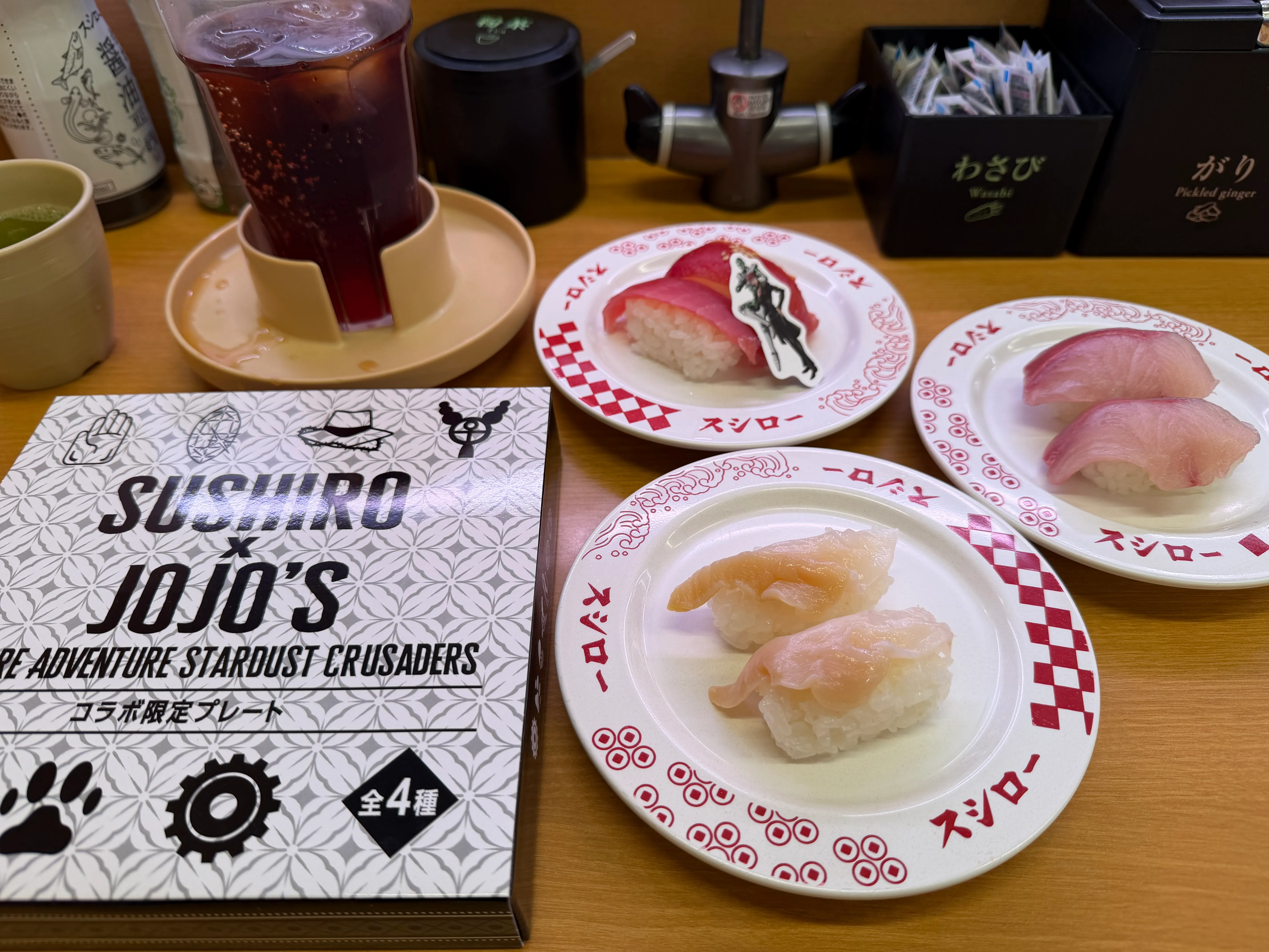 SUSHIRO × JOJO&#x27;S BIZARRE ADVENTURE STARDUST CRUSADERSのコラボ限定プレートの箱と、寿司3皿・ドリンクが並んだテーブル